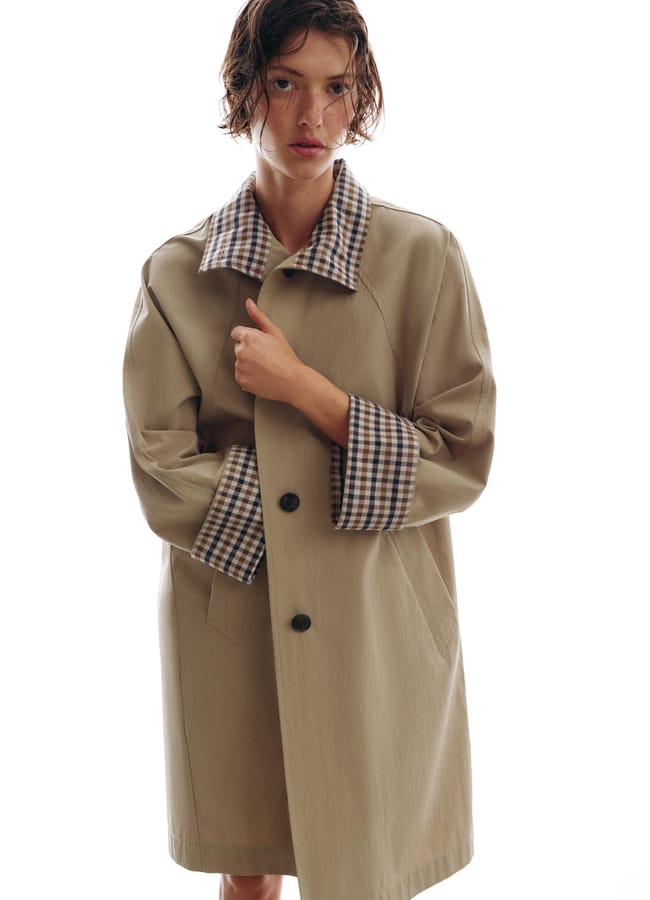 A person with short, wet hair wears one of the best trench coats—a beige style with a checked collar and cuffs—standing against a plain white background. They have a neutral expression and hold the coat closed with one hand.