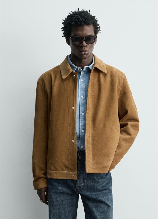 A person wearing dark sunglasses, a light blue button-up shirt, a tan men's jacket, and dark blue jeans stands against a plain light grey background.