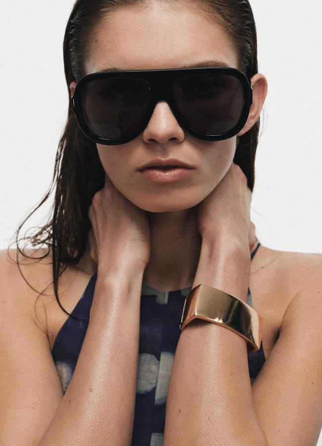 A woman with wet hair wears oversized black sunglasses, a sleeveless patterned top, and a wide gold cuff bracelet, holding her hands to her neck against a plain white background.