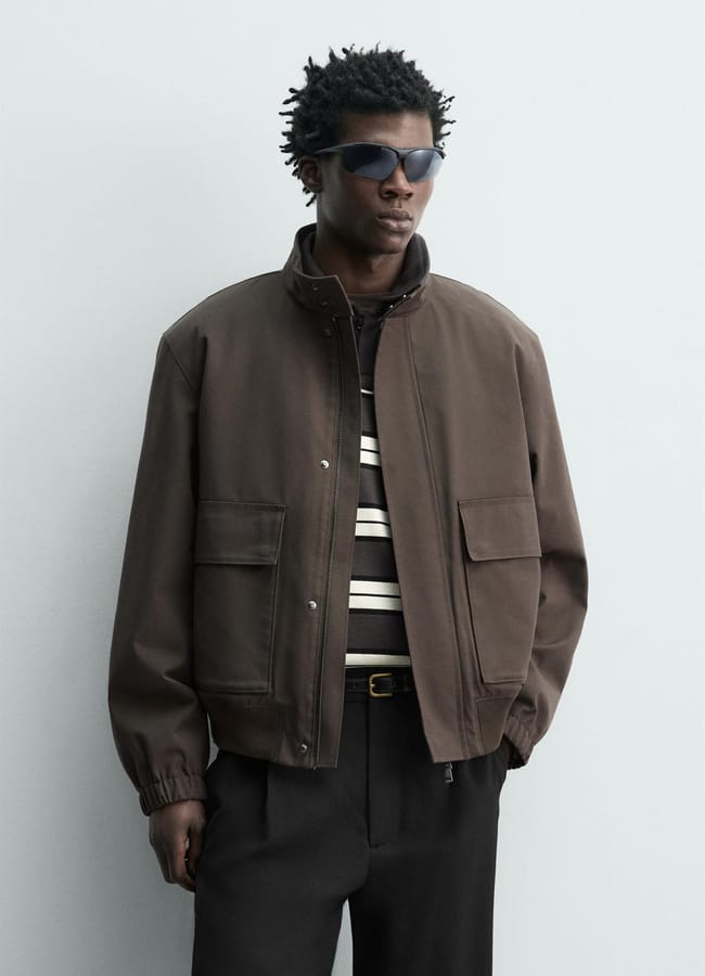 A man wearing dark sunglasses, a brown men's jacket over a striped shirt, and black trousers poses against a plain light grey background.