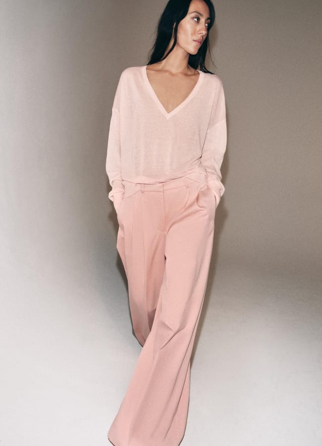 A woman with long dark hair wears a loose, light pink V-neck jumper and wide-legged, high-waisted pastel pink trousers—a chic look inspired by spring summer 2026 fashion trends—standing against a neutral background with hands in her pockets.