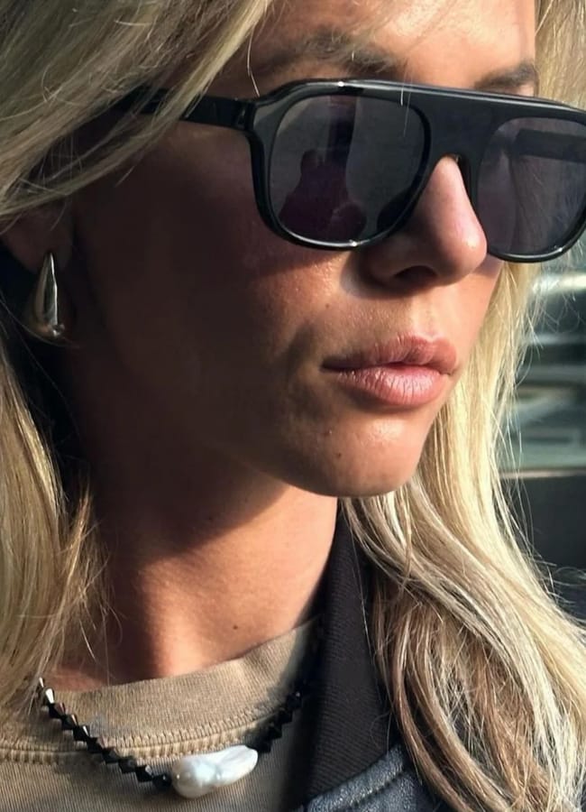A woman with long blonde hair wears large black sunglasses, a drop-shaped earring, and a necklace with a white stone. Sunlight highlights parts of her face and hair as she looks off to the side.