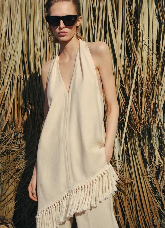 A woman wearing black sunglasses and a sleeveless, beige halter dress with fringe detail stands in front of tall, dry grass or reeds, one hand in her pocket, confidently channelling the allure of stylish fringe benefits.