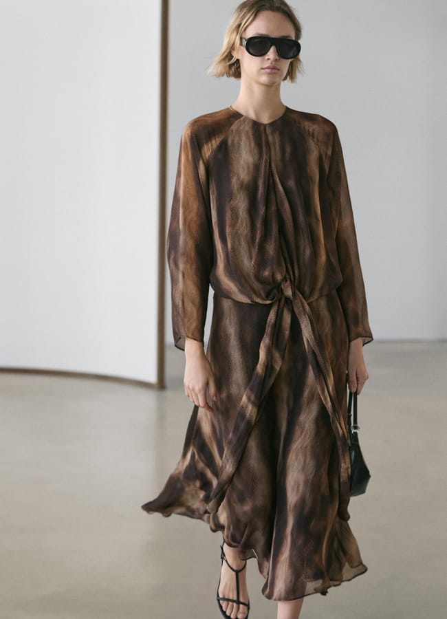 A woman wearing dark sunglasses and a flowing brown patterned wedding guest dress walks indoors holding a small black handbag. She has short hair, strappy black sandals, and the minimal background features white and wood panels.