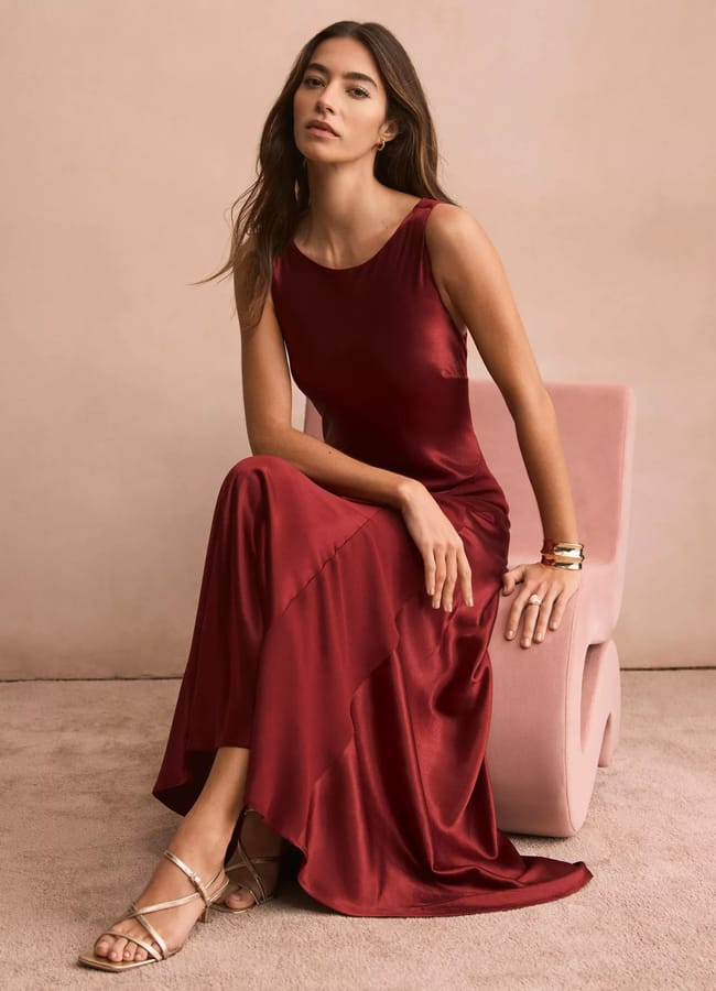 A woman in a sleeveless, deep red satin dress—one of our stunning wedding guest dresses—sits on a pale pink chair, looking at the camera. She has long brown hair, gold bracelets, and strappy sandals against a soft beige background.