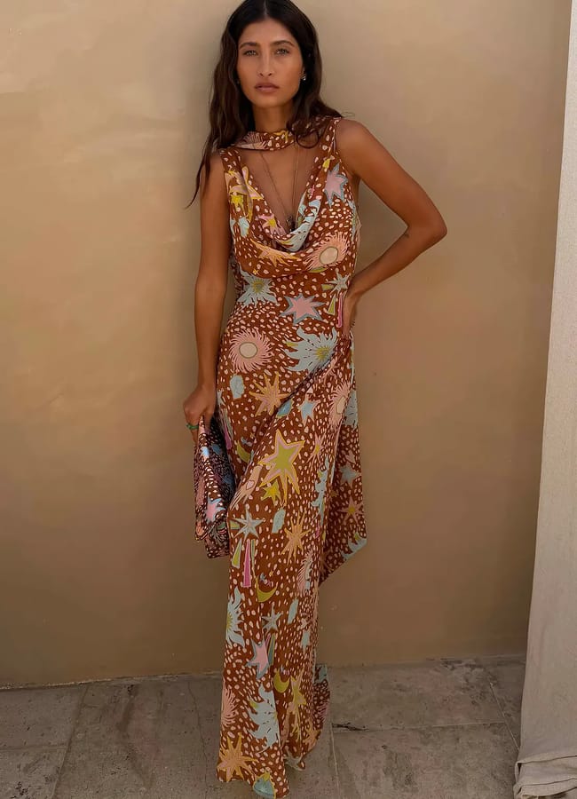 A woman stands against a beige wall wearing a sleeveless, floor-length dress with a colourful, abstract sun and star pattern—an elegant choice for wedding guest dresses. She holds a small clutch and gazes at the camera with a relaxed expression.