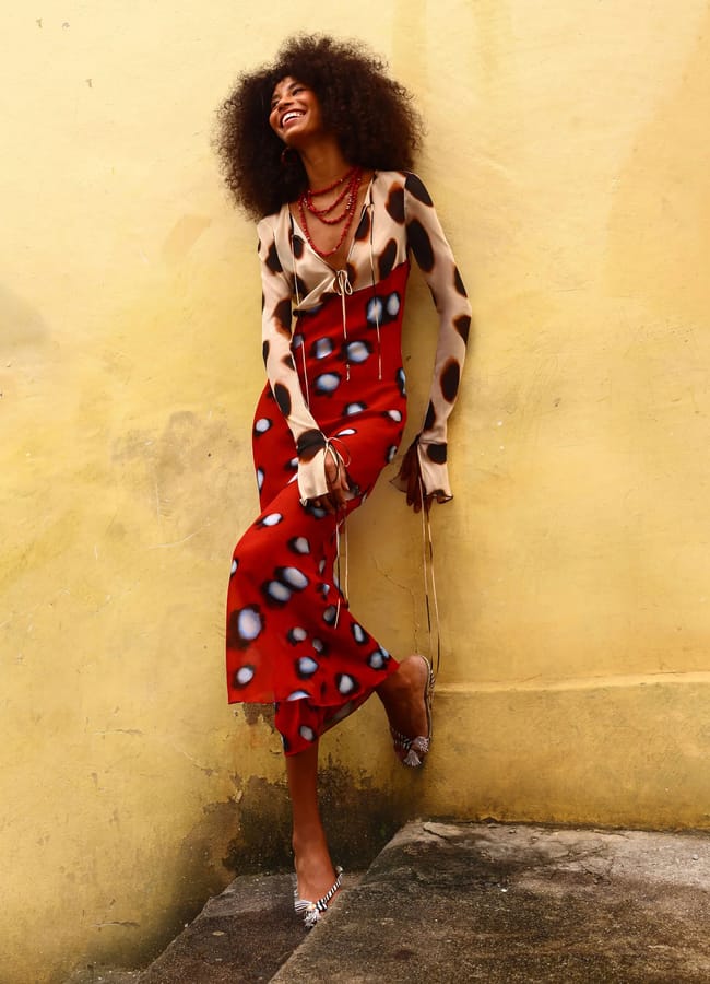 A woman with curly hair, smiling and leaning against a yellow wall, wears a fitted, patterned long-sleeve top and a red skirt with black and white spots—an eye-catching look perfect for those seeking unique wedding guest dresses.