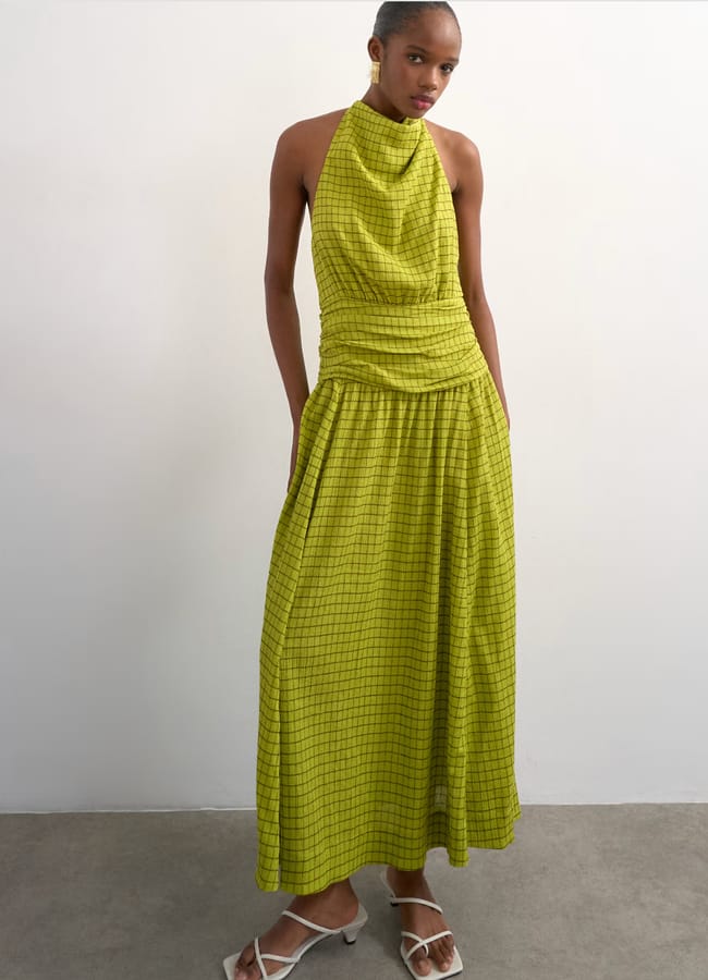 A woman stands against a light wall wearing a sleeveless, lime green chequered maxi dress with a halter neck and gathered waist—an elegant choice for wedding guest dresses. She completes the look with white strappy sandals.