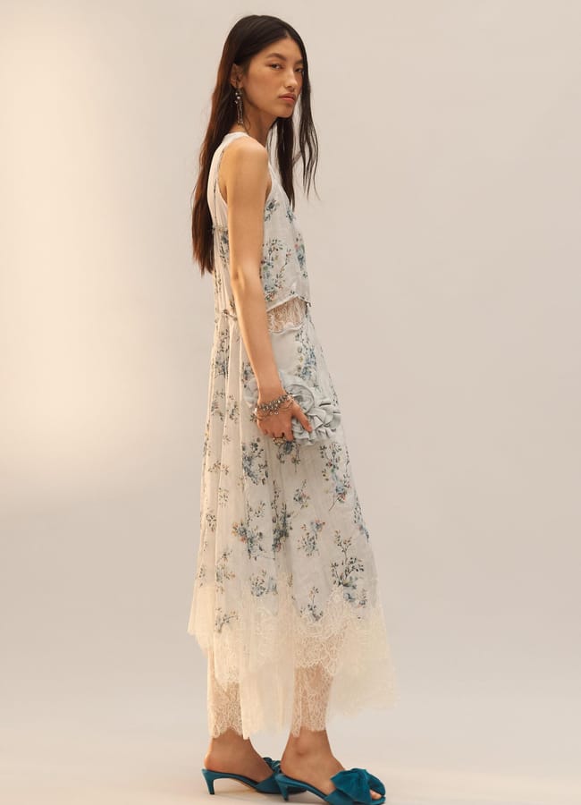 A woman with long dark hair stands sideways, wearing a sleeveless white lace dress with a floral pattern—perfect for wedding guest dresses—blue earrings, and teal open-toe mule heels. She holds a small clutch against a neutral background.