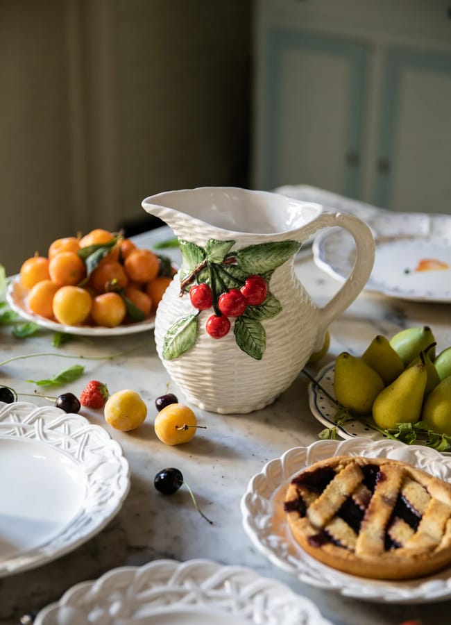 A ceramic jug with a cherry design sits on a table surrounded by plates of fresh fruit, including pears, cherries, and peaches, and a pie. Ornate white plates are set on the marble worktop.