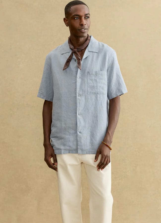 A man wearing a light blue short-sleeve button-up shirt, cream trousers, and a brown scarf tied around his neck stands against a beige background, showcasing relaxed men's holiday fashion as he looks slightly to the side.