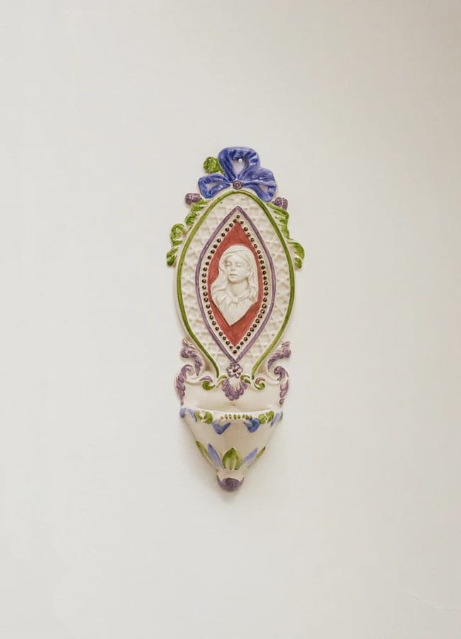 Ornate ceramic wall plaque with a red-bordered cameo of a woman’s profile, surrounded by green, blue, and purple floral decorations, and a small cup or holder at the bottom.