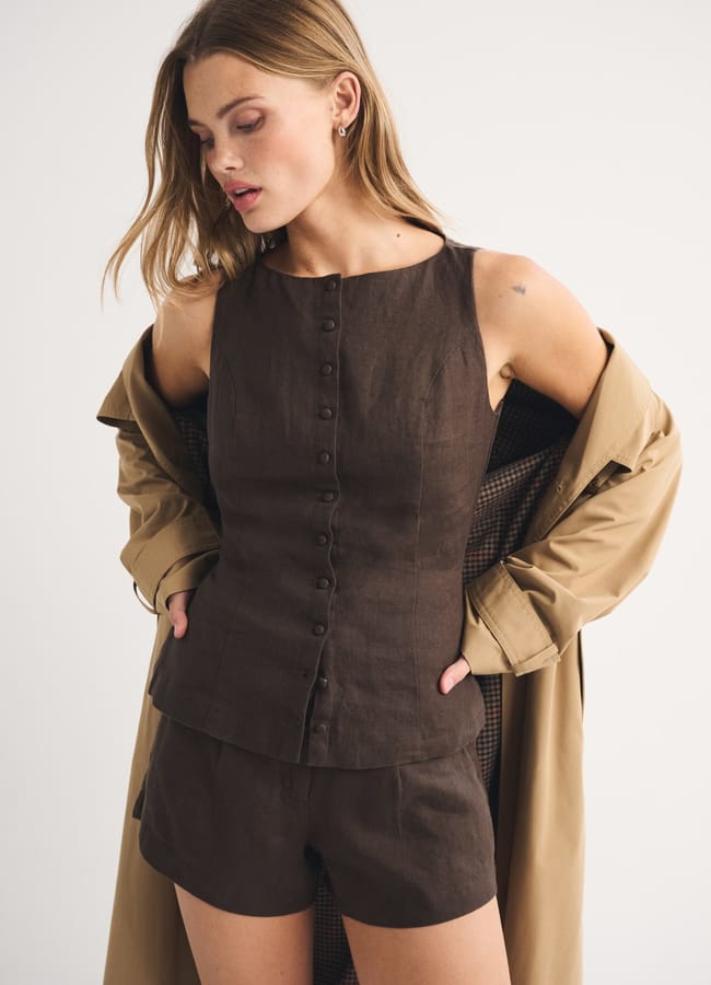 Woman wearing a sleeveless, button-up brown top and matching shorts, with a beige high street linen trench coat draped over her shoulders, standing against a plain white background.