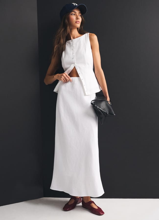 A woman stands against a black-and-white wall, wearing a high street linen sleeveless white top with front buttons, a long white skirt, burgundy shoes, a navy baseball cap, and holding a black clutch bag.