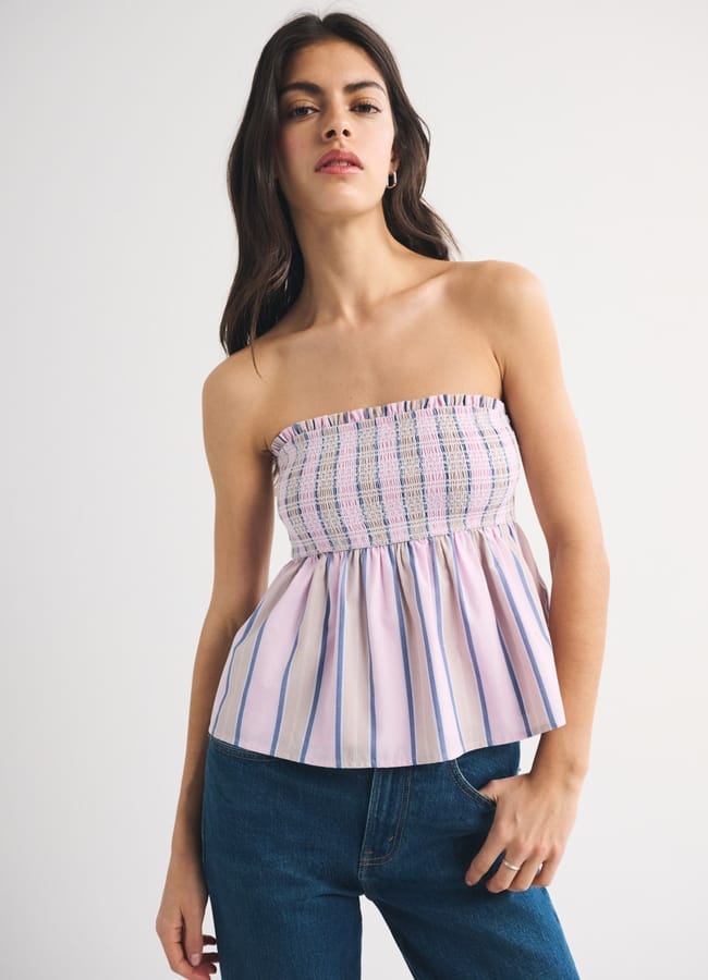 A woman with long brown hair wears a strapless, ruffled summer top featuring a shirred, multicoloured upper section and vertical blue and pink stripes below, paired with blue jeans. She stands against a plain light background.