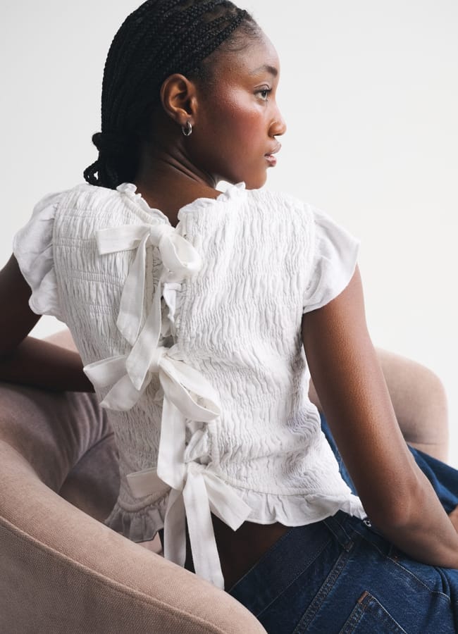A woman with plaited hair sits sideways in a light-coloured chair, wearing a white summer top with ribbon ties down the back and blue jeans, looking off to the side against a plain background.