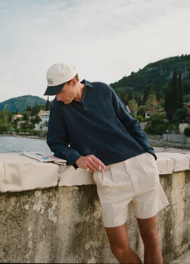 A person showcases men's holiday fashion in a navy long-sleeve shirt, cream shorts, and a baseball cap while leaning against a stone wall by the water, with hills and houses in the background under a partly cloudy sky.