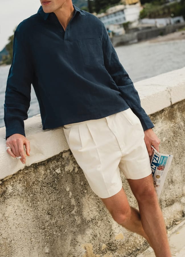 A man in a navy long-sleeve shirt and cream pleated shorts—capturing effortless men's holiday style—leans against a stone wall by the water, cigarette in one hand and a rolled-up magazine in the other; houses and trees line the background.