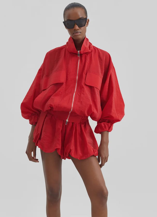 A person wearing sunglasses poses in a bright red, loose-fitting zip-up jacket with large front pockets and matching red bubble shorts against a plain light grey background.