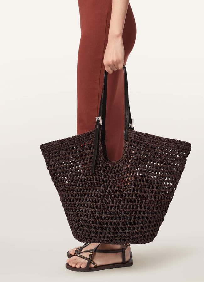 A person in a rust-coloured dress and black sandals holds a large, dark brown woven basket bag with black handles against a plain light background.
