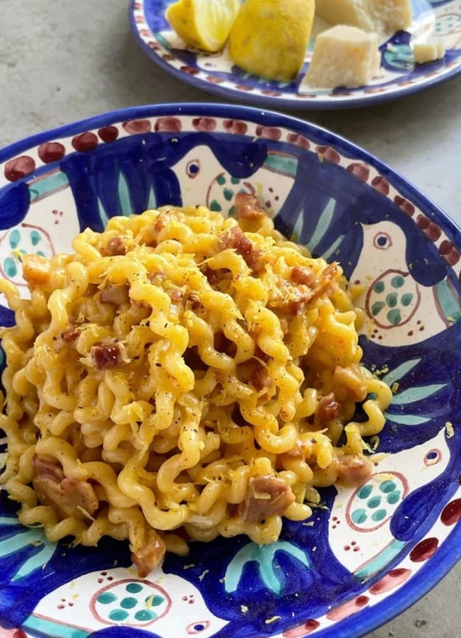 A colourful ceramic plate holds a serving of curly pasta mixed with bits of bacon and black pepper. In the background, a plate with lemon wedges and cheese is partially visible.