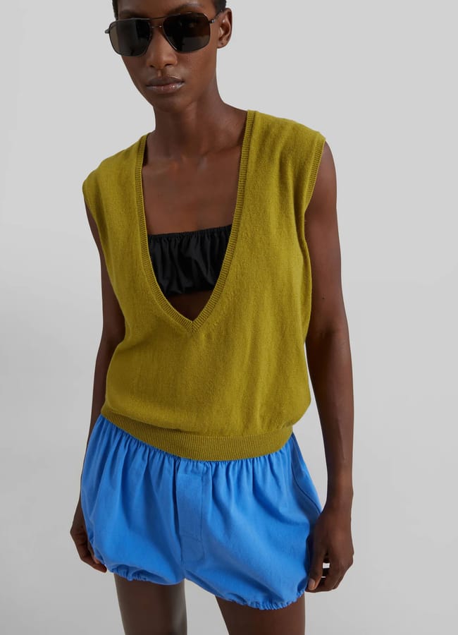 A person wearing dark sunglasses, a sleeveless deep V-neck olive green jumper over a black bandeau top, and bright blue bubble shorts stands against a plain light grey background.