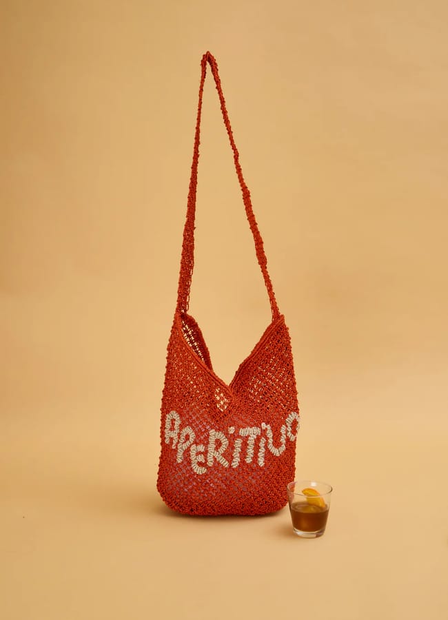 A red knitted shoulder bag, inspired by classic basket bags, with the word APERITIVO in white letters sits on a beige surface next to a glass of brown liquid with an orange slice. The background is a plain beige colour.
