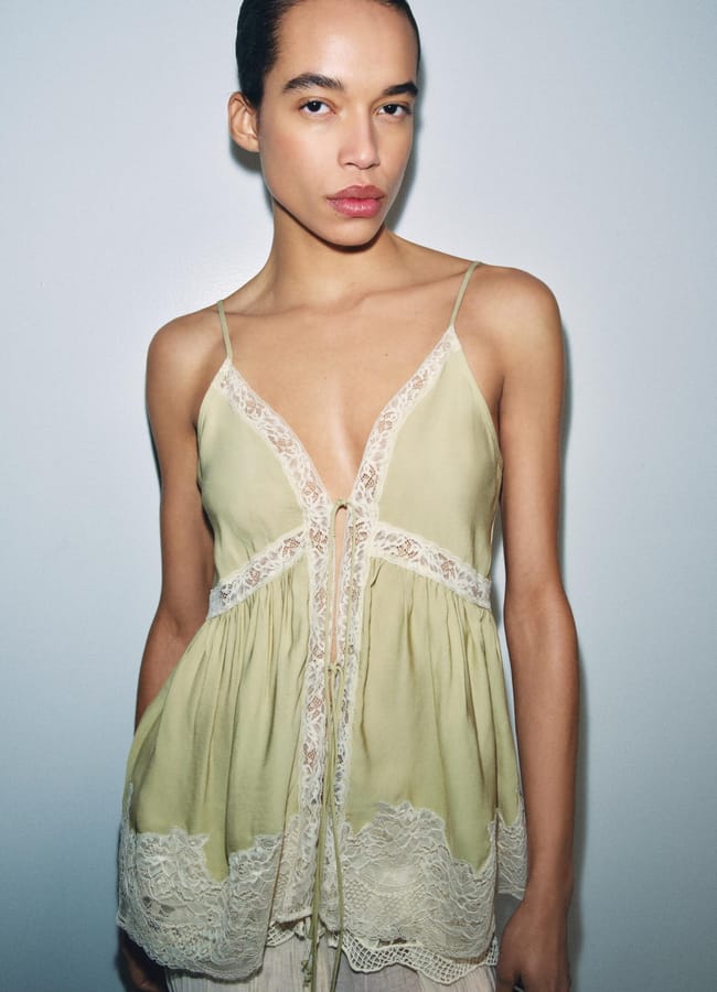 A person with dark hair pulled back wears a light green, lace-trimmed camisole—one of this season's must-have summer tops—featuring thin straps and a deep neckline, standing against a plain light blue background.