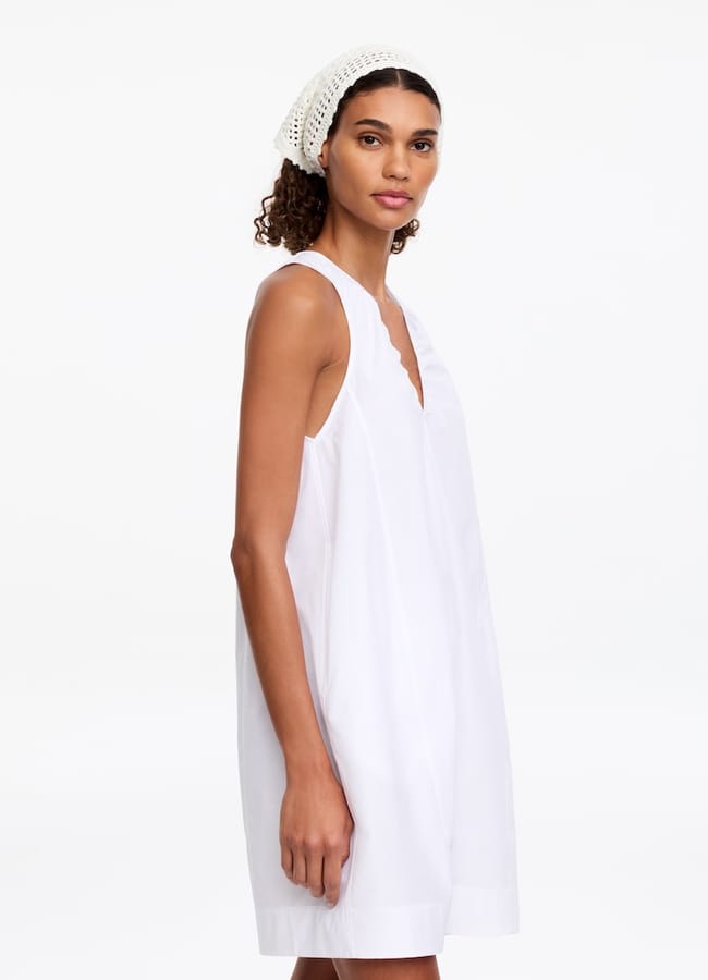 A woman wearing a sleeveless, loose-fitting white dress and a white crochet headband stands against a plain white background, looking slightly to the side.