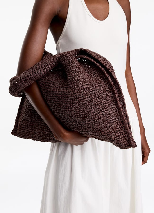 A person in a sleeveless white dress holds a large, textured brown basket bag under their arm against a plain white background.
