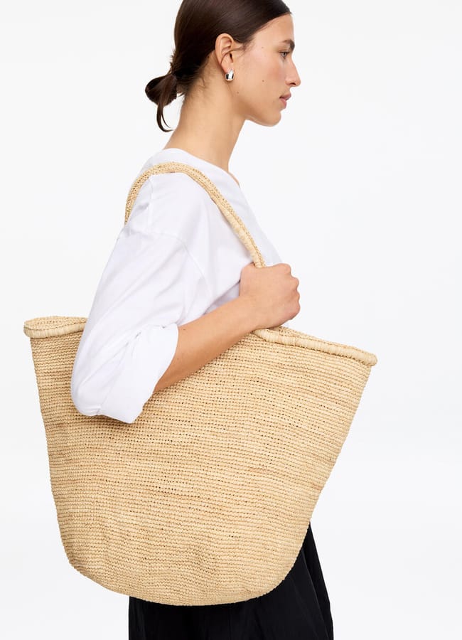 A woman in a white shirt and black trousers carries a large, woven straw basket bag over her shoulder against a plain white background.