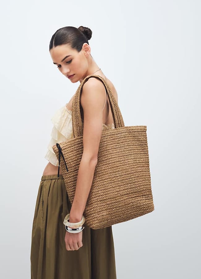 A woman with dark hair in a bun wears a cream ruffled top and olive green skirt, holding a large woven basket bag over her shoulder against a plain light background.