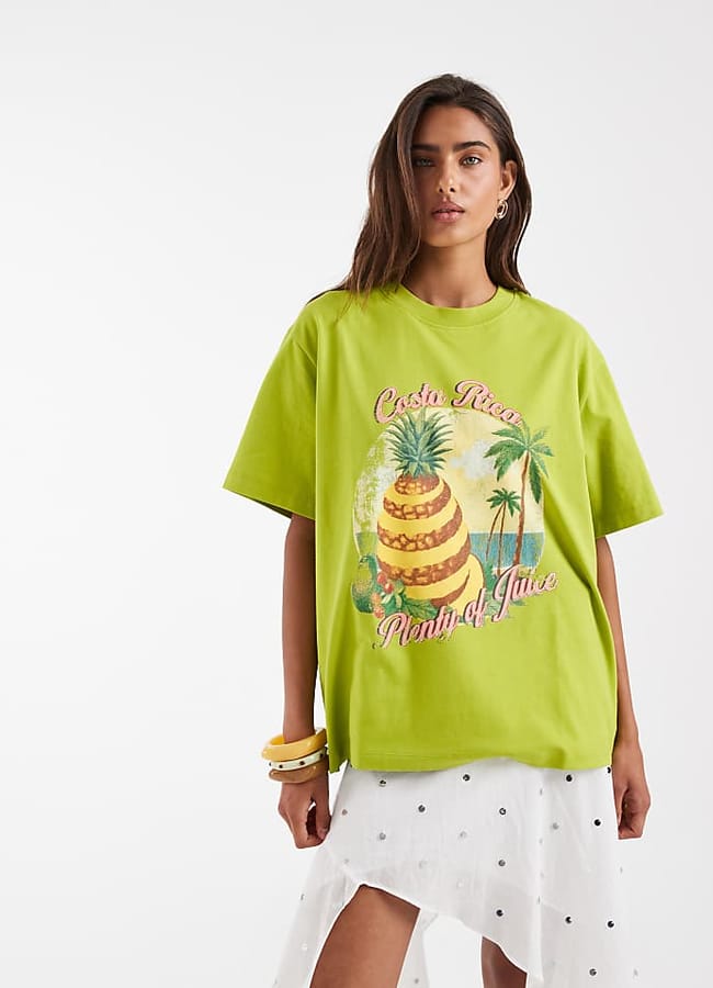 A woman in a lime green Costa Rica Plenty of Juice T-shirt with a pineapple graphic and white skirt with black polka dots stands against a plain white background, showing off her love for fruit prints. She wears yellow and white bracelets.