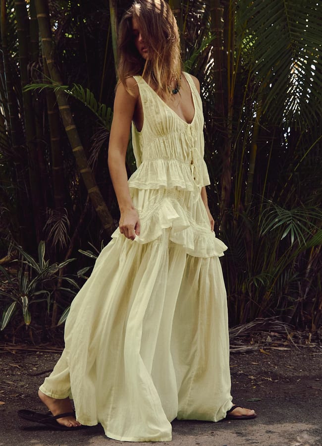 A woman stands outdoors among lush green plants, wearing a flowing, sleeveless, light yellow ruffled maxi dress and black sandals—an effortlessly stylish look that could inspire unique Father’s Day gifts 2026. Her long hair partially covers her face.