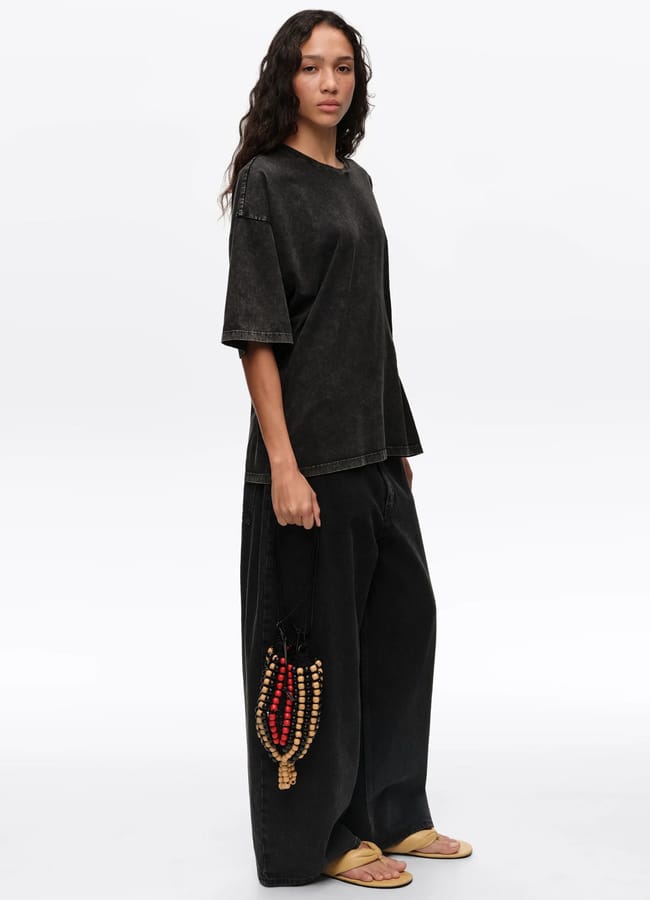 A woman wearing a loose-fitting, faded black T-shirt and wide-leg trousers stands against a plain background, holding a stylish beaded bag and wearing flip-flops.