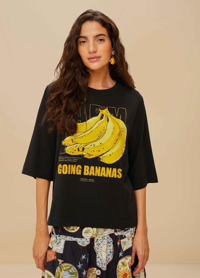 A woman with long wavy brown hair wears a black T-shirt featuring large yellow bananas and the words GOING BANANAS, paired with a patterned skirt adorned with fruit prints and decorative motifs, standing against a plain beige background.