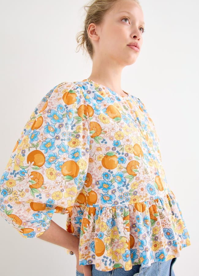 A person stands against a plain white background, wearing a loose, colourful blouse with vibrant fruit prints and a pattern of blue, yellow, and orange flowers. The blouse features puffy sleeves and a ruffled hem. The person looks upwards, hair tied back.