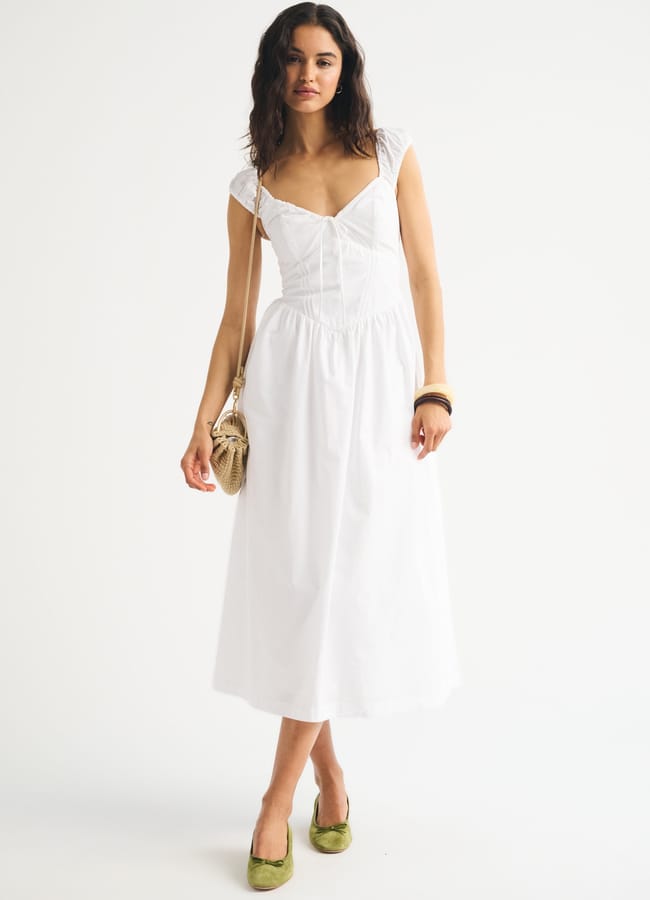 Woman in a white sleeveless midi dress with lace details, holding a tan woven handbag, wearing green flat shoes and wooden bangles, standing against a plain white background.