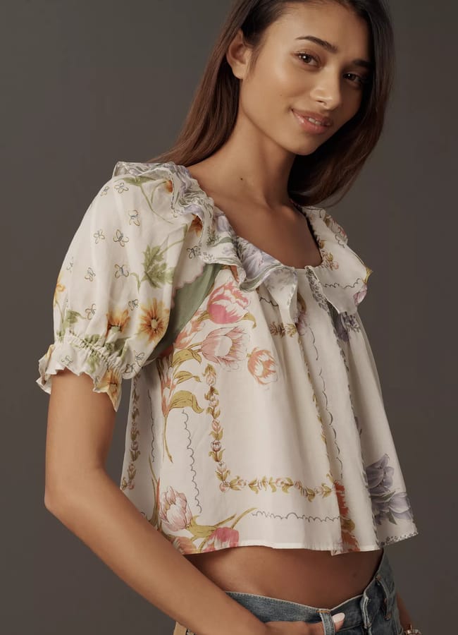 A woman with long brown hair wears a short-sleeved, ruffled, floral blouse in pastel colours—a perfect choice for summer tops. She stands against a plain, dark background and smiles slightly whilst looking at the camera.