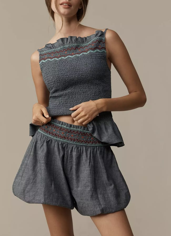 A woman models a sleeveless, ruffled, smocked blue-grey top with matching high-waisted bubble shorts featuring embroidered details against a plain beige background. Her face is partially out of shot.