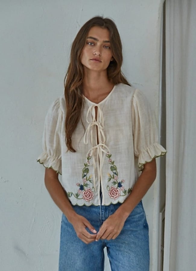 A woman with long brown hair wears a cream-coloured, short-sleeved blouse with floral embroidery—one of the season’s must-have summer tops—and blue jeans. She stands against a light-coloured wall with a neutral expression.