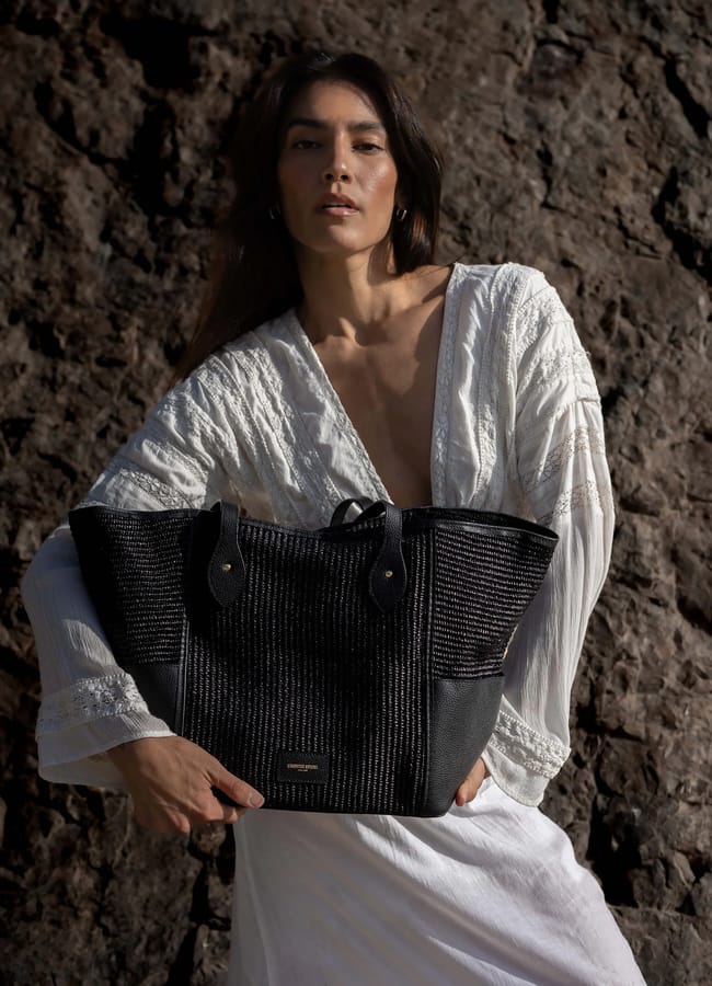 A woman in a white, long-sleeve, deep V-neck dress stands outdoors against a rocky background, holding a large black textured tote bag with gold hardware and handles—a chic take on classic basket bags.