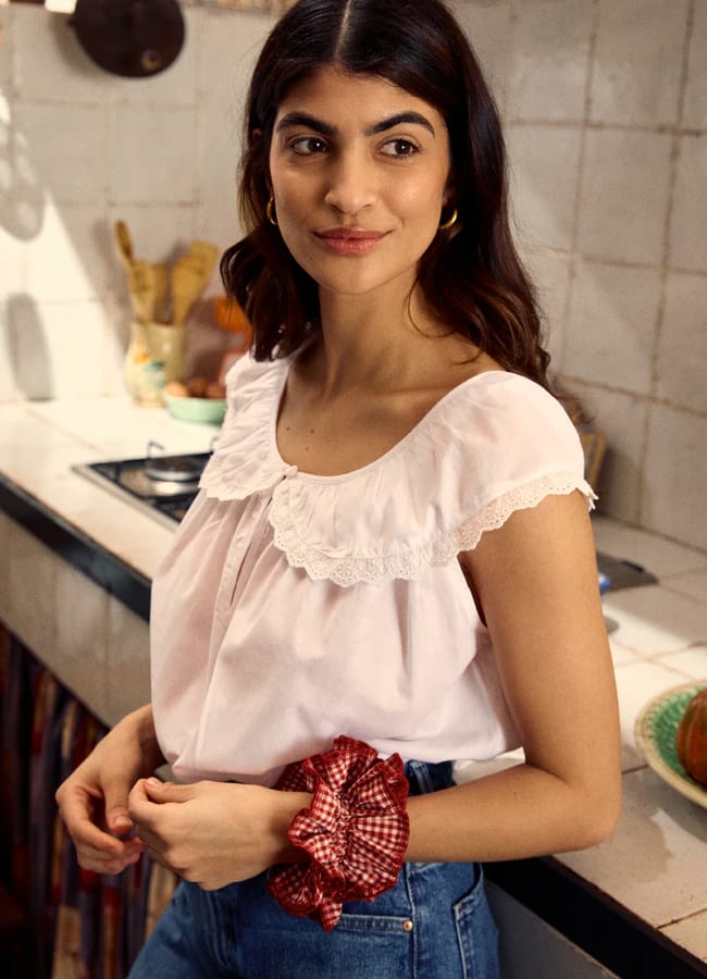 A woman with long brown hair, wearing a white summer top with a lace collar and blue jeans, stands in a cosy kitchen, holding a red gingham scrunchie and smiling slightly.