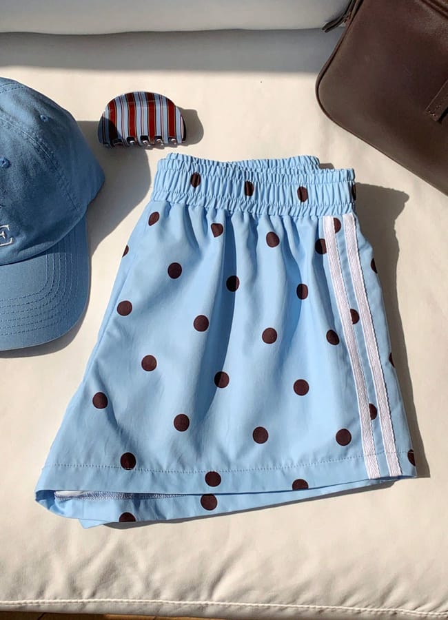 A light blue skirt with a brown polka dot pattern, white side stripes, and an elasticated waistband—perfect for cute padel outfits—is displayed next to a blue baseball cap, a striped hair clip, and a brown bag on a cream surface.