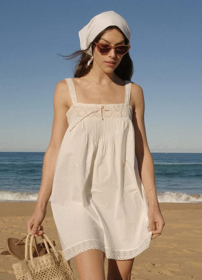 A woman embracing nonnamaxxing style in a white sundress, headscarf, and sunglasses strolls along a sandy beach with a woven bag, set against the sea and clear blue sky.