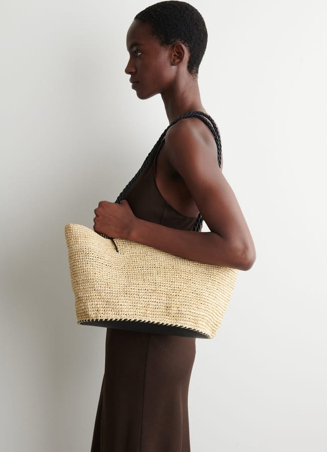 A person in a sleeveless brown dress stands in profile against a white wall, holding a woven straw basket bag with black handles over their shoulder.