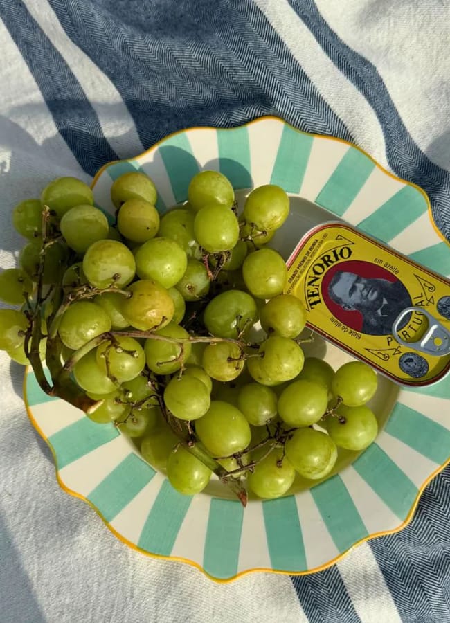 A striped plate with green grapes and an open tin of Tenorio sardines rests on a white and blue striped fabric in sunlight.