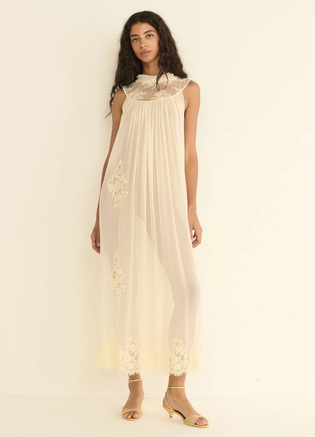 A woman with long curly hair wears a sheer, cream-coloured, floor-length dress with lace details and beige heeled sandals, standing against a plain off-white background—an elegant look perfect for summer or special occasions like Father’s Day gifts 2026.