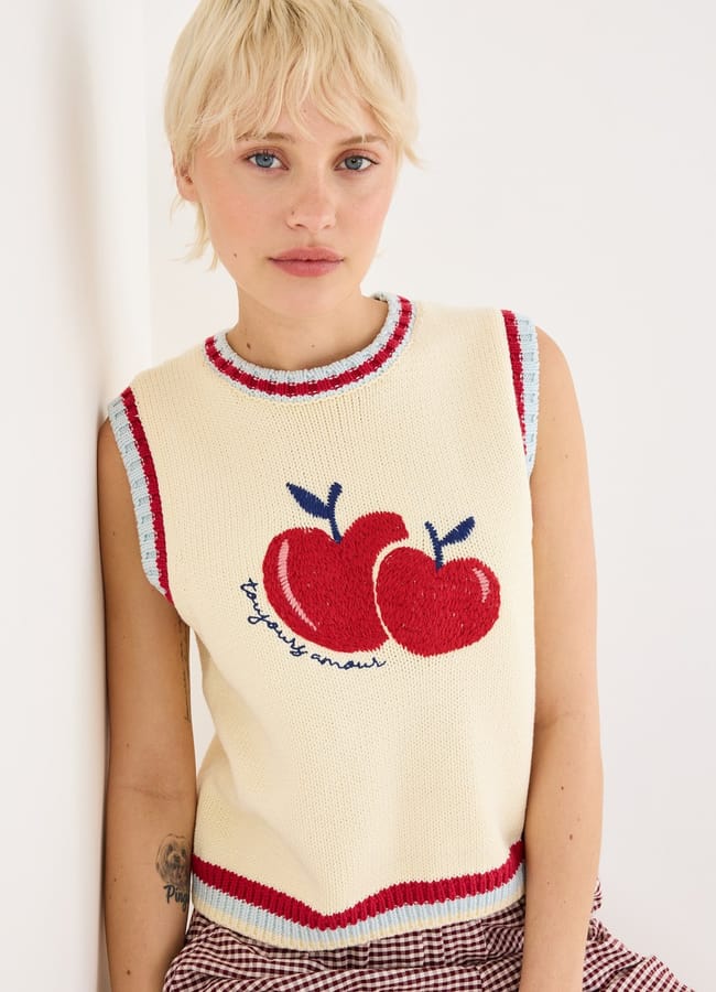 A person with short blond hair wears a sleeveless cream jumper vest with fruit prints—two red apples and the text “for juicy ones!”—alongside checked bottoms. They lean against a white wall, blending playful style with bold details.