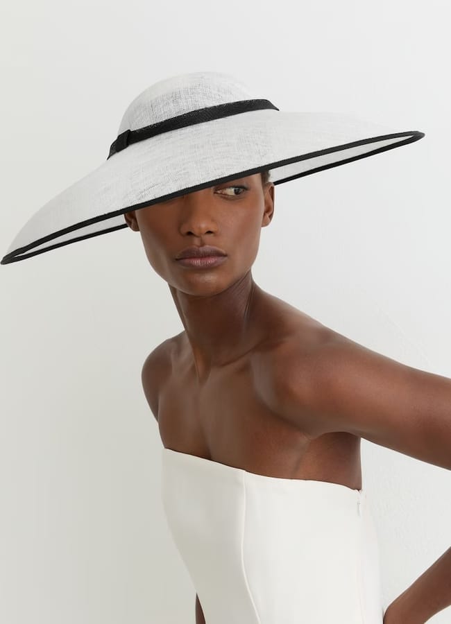 A woman wearing a strapless white dress and a wide-brimmed, white hat with a black band—clearly a bold take on banned fascinators—poses against a plain white background, the hat’s shadow partially obscuring her face.